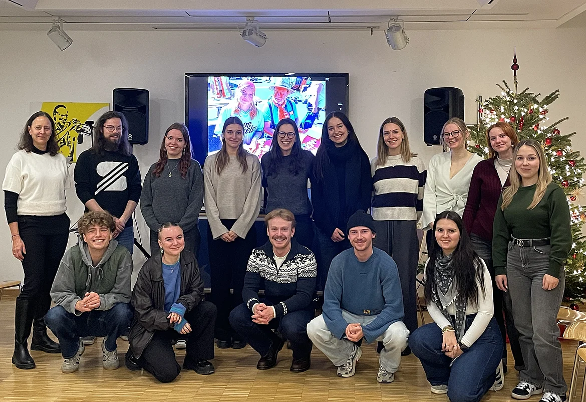 Eine Gruppe von 15 Personen posiert in einem hellen Raum vor einem Bildschirm. Die Gruppe besteht aus jüngeren und älteren Menschen, einige sitzen auf dem Boden, andere stehen dahinter. Alle blicken freundlich in die Kamera und wirken entspannt und gut gelaunt. Im Hintergrund sind weiße Wände, ein geschmückter Weihnachtsbaum und technische Geräte zu sehen.