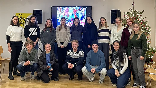 Eine Gruppe von 15 Personen posiert in einem hellen Raum vor einem Bildschirm. Die Gruppe besteht aus jüngeren und älteren Menschen, einige sitzen auf dem Boden, andere stehen dahinter. Alle blicken freundlich in die Kamera und wirken entspannt und gut gelaunt. Im Hintergrund sind weiße Wände, ein geschmückter Weihnachtsbaum und technische Geräte zu sehen.