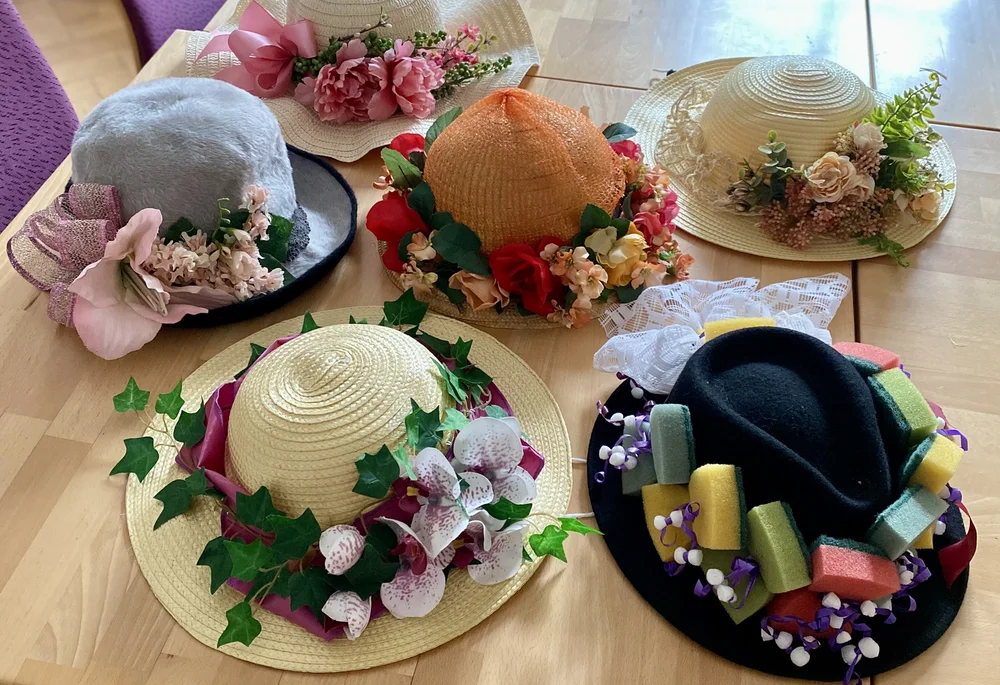 Das Bild zeigt eine Auswahl an dekorativen Hüten, die mit Blumen, Blättern, Bändern und anderen Verzierungen geschmückt sind. Die Hüte haben verschiedene Farben, darunter grau, beige und orange. Sie liegen auf einem Tisch und sind Teil des kreativen Projekts "Verrückte Hüte".