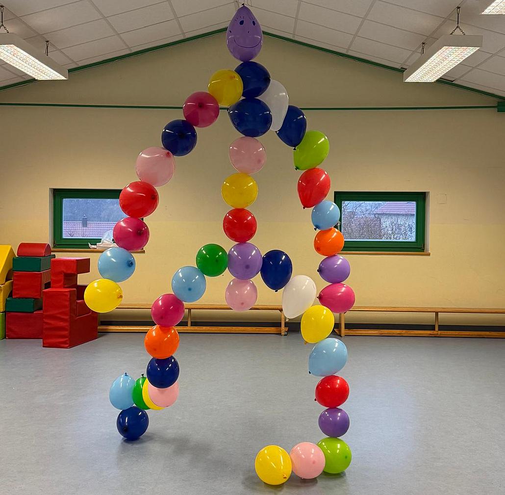 Das Bild zeigt einen großen Raum, eine Turnhalle, mit hellem Boden, cremefarbenen Wänden und grünen Fensterrahmen. In der Mitte des Raumes steht eine Figur aus vielen bunten Luftballons. Die Ballons sind in verschiedenen Farben wie Rot, Blau, Gelb, Grün, Rosa, Lila und Weiß. Sie sind so angeordnet, dass sie die Form eines menschlichen Körpers bilden: zwei Beine, ein Oberkörper, zwei Arme und ein Kopf.