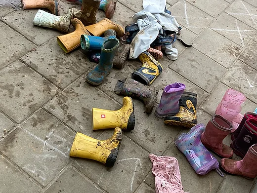 Das Bild zeigt viele bunte Kindergummistiefel voll mit Matsch auf Asphalt liegend. Dazwischen sind auch zwei sehr dreckige Matschhosen zu sehen. 