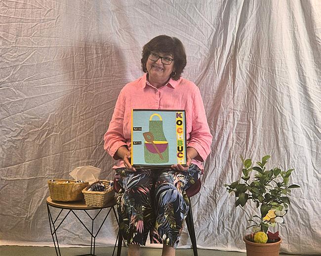Eine Frau sitzt auf einem Stuhl vor einer weißen Wand, sie lächelt und hält einen Ordner mit der Aufschrift „Kochbuch“ in der Hand. Rechts neben ihr stehen auf einem kleinen Tisch zwei Körbe, links von ihr ein kleiner Zitronenbaum.