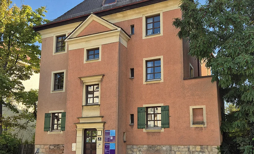 Abgebildet ist ein dreigeschossiges, rosafarbenes Gebäude mit klassischer Fassade, Satteldach und grünen Fensterläden, umgeben von Bäumen. Im unteren Bereich des Bildes befindet sich ein dunkelblauer Balken mit der Aufschrift „Beratungen & Hilfe – Sozialpsychiatrischer Dienst“. 