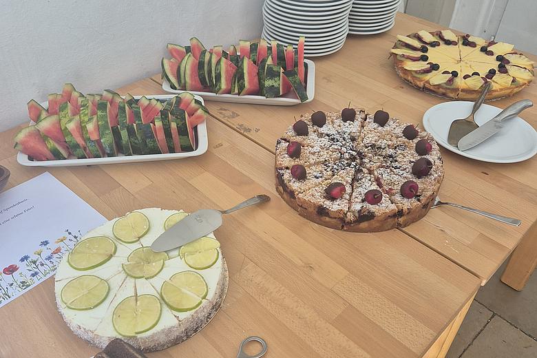 Das Bild zeigt einen Tisch mit verschiedenen Kuchen in Buffetform angerichtet. Im hinteren Bereich sind zwei Platten mit Wassermelonenstücke. In der Ecke rechts oben sind Teller gestapelt. 