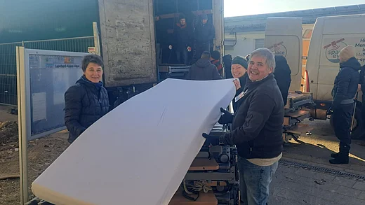 Mehrere Menschen laden gemeinsam Pflegebetten und Matratzen in einen LKW. Im Vordergrund tragen zwei Helferinnen lächelnd eine weiße Matratze. Im Hintergrund sind weitere Helferinnen und Helfer sowie ein geöffneter LKW zu sehen. Daneben stehen Transportfahrzeuge der Hilfsorganisation Space-Eye. Die Szene zeigt die Verladung der gespendeten Pflegeausstattung vor dem Leonhard-Kaiser-Haus.
