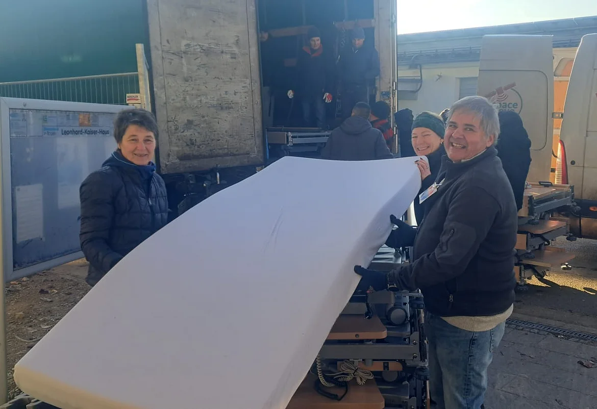 Mehrere Menschen laden gemeinsam Pflegebetten und Matratzen in einen LKW. Im Vordergrund tragen zwei Helferinnen lächelnd eine weiße Matratze. Im Hintergrund sind weitere Helferinnen und Helfer sowie ein geöffneter LKW zu sehen. Daneben stehen Transportfahrzeuge der Hilfsorganisation Space-Eye. Die Szene zeigt die Verladung der gespendeten Pflegeausstattung vor dem Leonhard-Kaiser-Haus.