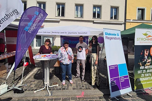 Menschen stehen an einem Informationsstand der Diakonie Regensburg auf einem gepflasterten Platz in der Innenstadt. Vor einem weißen Pavillon mit Fahnen und Roll-ups informieren Mitarbeitende über Angebote zur psychischen Gesundheit und soziale Unterstützung. Im Hintergrund sind historische Gebäude bei sonnigem Wetter zu sehen.