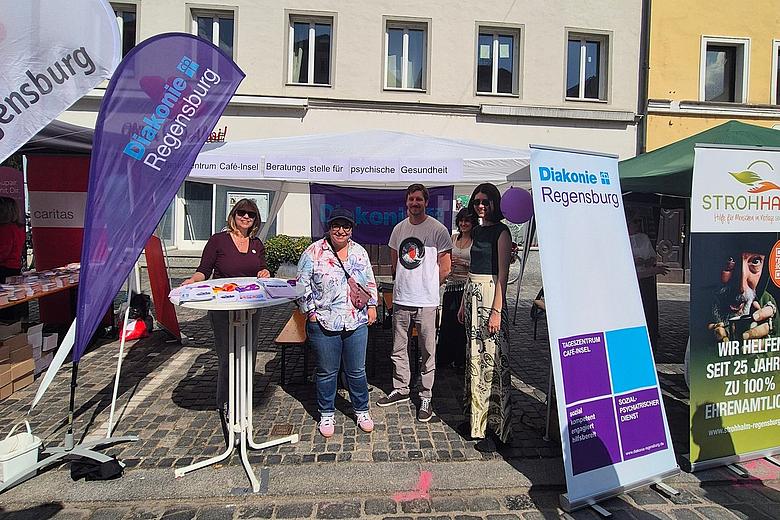 Menschen stehen an einem Informationsstand der Diakonie Regensburg auf einem gepflasterten Platz in der Innenstadt. Vor einem weißen Pavillon mit Fahnen und Roll-ups informieren Mitarbeitende über Angebote zur psychischen Gesundheit und soziale Unterstützung. Im Hintergrund sind historische Gebäude bei sonnigem Wetter zu sehen.