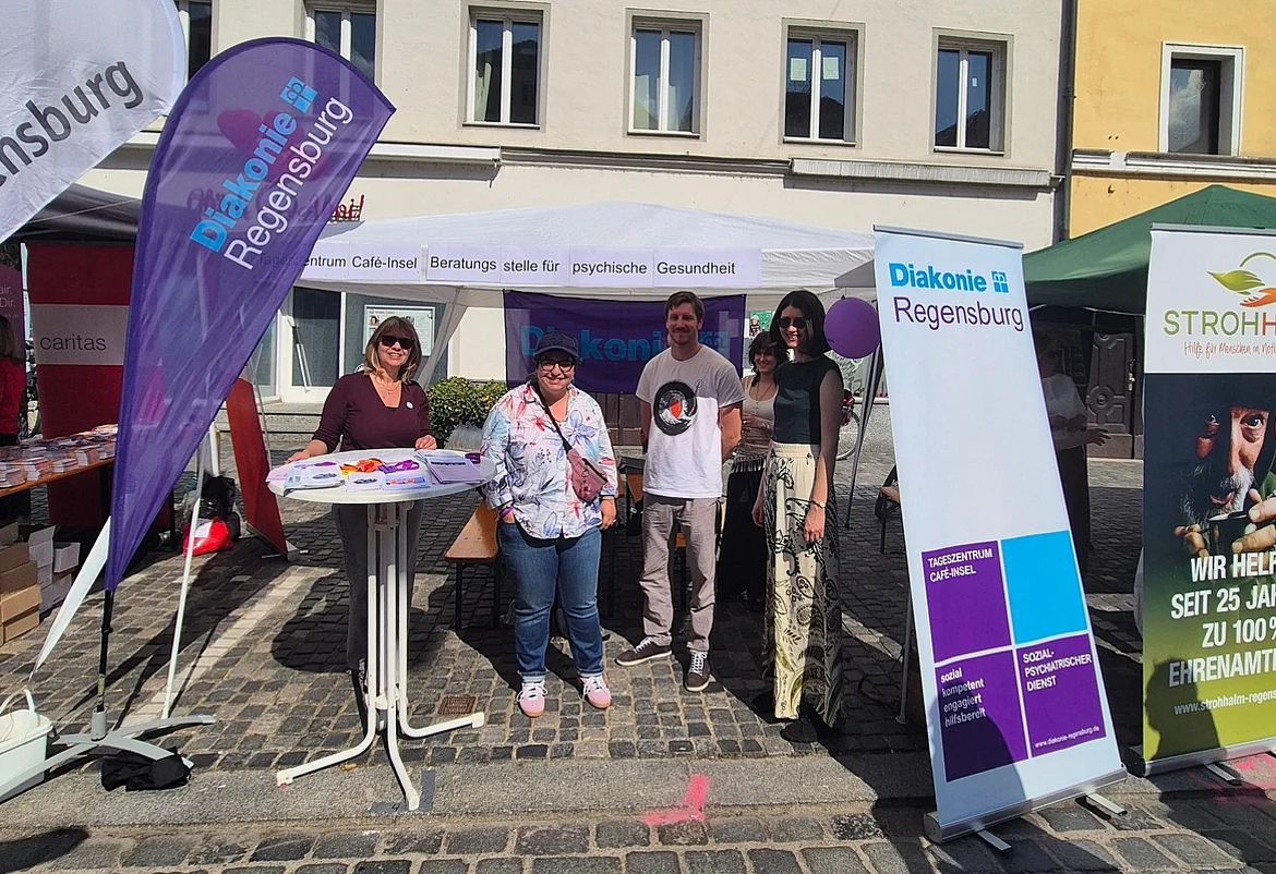 Menschen stehen an einem Informationsstand der Diakonie Regensburg auf einem gepflasterten Platz in der Innenstadt. Vor einem weißen Pavillon mit Fahnen und Roll-ups informieren Mitarbeitende über Angebote zur psychischen Gesundheit und soziale Unterstützung. Im Hintergrund sind historische Gebäude bei sonnigem Wetter zu sehen.