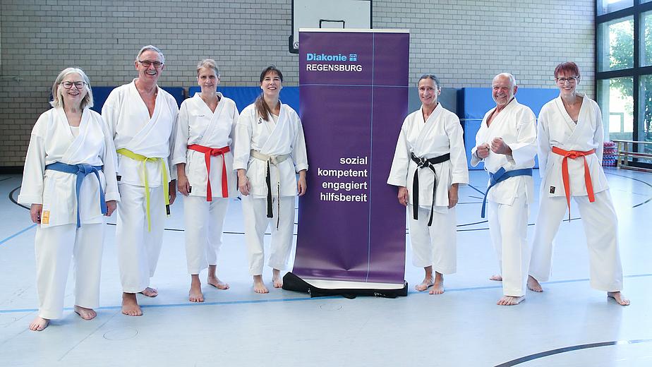 Sieben Menschen in weißer Karatekleidung stehen in einer Turnhalle. Sie tragen alle verschiedene Bunte Gürtel. In der Mitte steht ein lila Banner mit der Aufschrift: Diakonie Regensburg. sozial, kompetent, engagiert, hilfsbereit