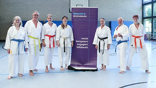 Sieben Menschen in weißer Karatekleidung stehen in einer Turnhalle. Sie tragen alle verschiedene Bunte Gürtel. In der Mitte steht ein lila Banner mit der Aufschrift: Diakonie Regensburg. sozial, kompetent, engagiert, hilfsbereit
