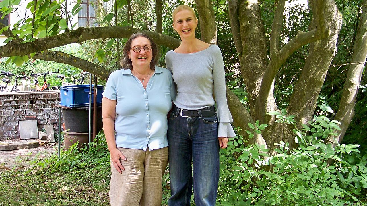 Zwei Frauen stehen nebeneinander im Freien in einem grünen Gartenbereich mit Bäumen und Sträuchern. Beide lächeln in die Kamera. Eine Frau trägt ein hellblaues Oberteil und eine helle Hose, die andere ein helles langärmeliges Oberteil und eine dunkle Jeans.