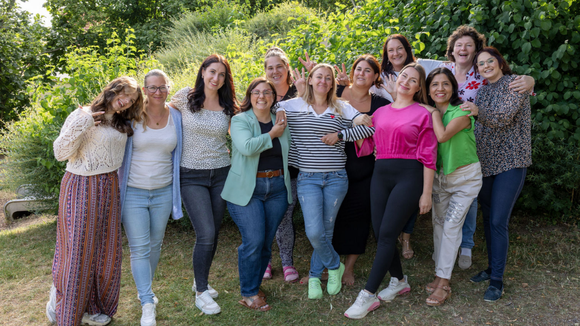 Das Bild zeigt 12 Frauen, die bunt gekleidet sind. Sie stehen eng zusammen, zum Teil in lustigen Posen. Im Hintergrund sind grüne Sträucher.