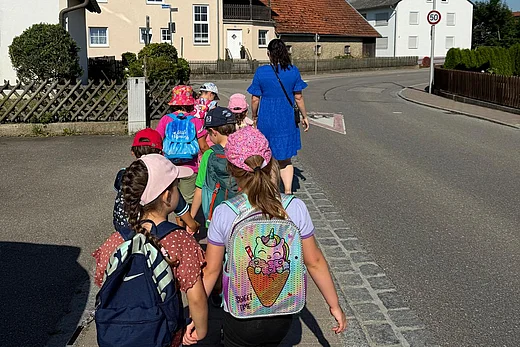 Das Bild zeigt eine Gruppe mit 7 Vorschulkindern eine Straße entlang gehen und von hinten zu sehen sind. Vorne geht eine Erzieherin. Die Kinder sind bunt gekleidet.