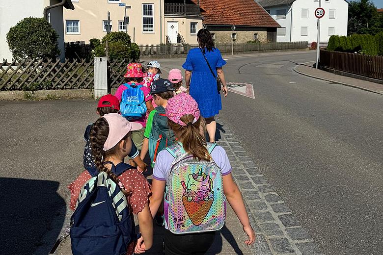 Das Bild zeigt eine Gruppe mit 7 Vorschulkindern eine Straße entlang gehen und von hinten zu sehen sind. Vorne geht eine Erzieherin. Die Kinder sind bunt gekleidet.