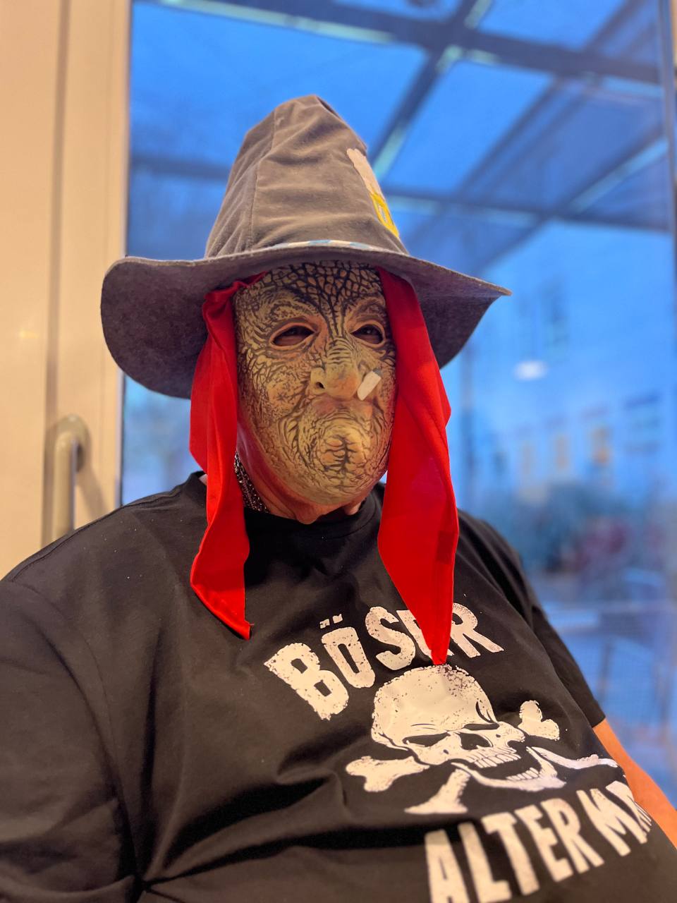 Das Bild wurde während der Faschingsfeier aufgenommen. Es zeigt eine Person, die eine Verkleidung trägt. Sie trägt eine Maske mit einem gealterten, runzeligen Gesicht, einen spitzen Hut und rote Stoffbänder statt Haare. Auf dem schwarzen T-Shirt steht die Aufschrift "Böser Alter" und ein Totenkopf. Im Hintergrund ist ein Fenster zu sehen.