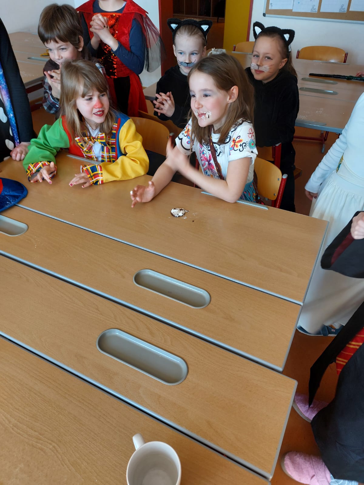 Das Bild zeigt eine Gruppe von Kindern des Kinderhortes Nittendorf bei der Faschingsfeier. Die Kinder sind verkleidet. Zwei Mädchen sitzen an einem Holztisch, während andere Kinder um die beiden herumstehen und lachen. Eines der Mädchen trägt ein buntes Kostüm, ähnlich einer Clownverkleidung. Das andere Mädchen trägt ein helles, gemustertes Oberteil. Im Hintergrund sind weitere Kinder zu sehen, einige mit Katzenohren und aufgemalten Schnurhaaren. Die Stimmung wirkt fröhlich. 